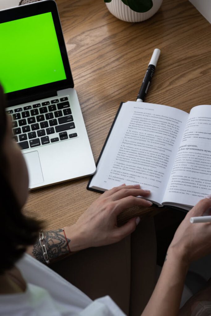 A woman studies at home using a laptop and an open book, focused on her homework.